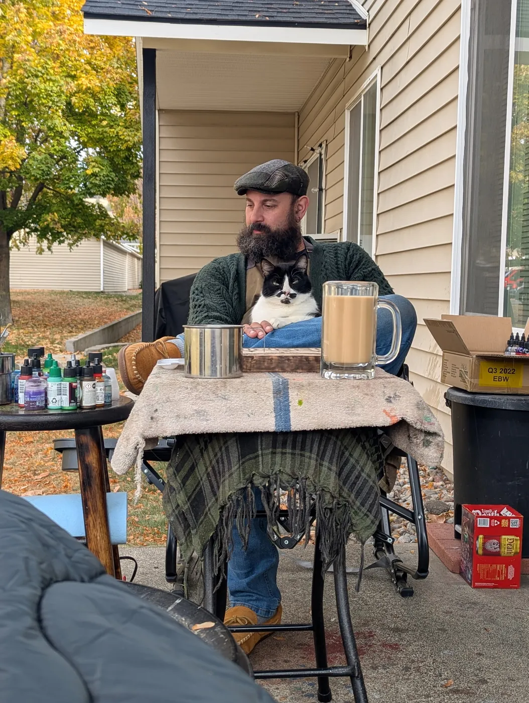 Tyler in a flat cap and green sweater at the patio workbench, Rorschach curled in his lap, paint bottles and two mugs on the table