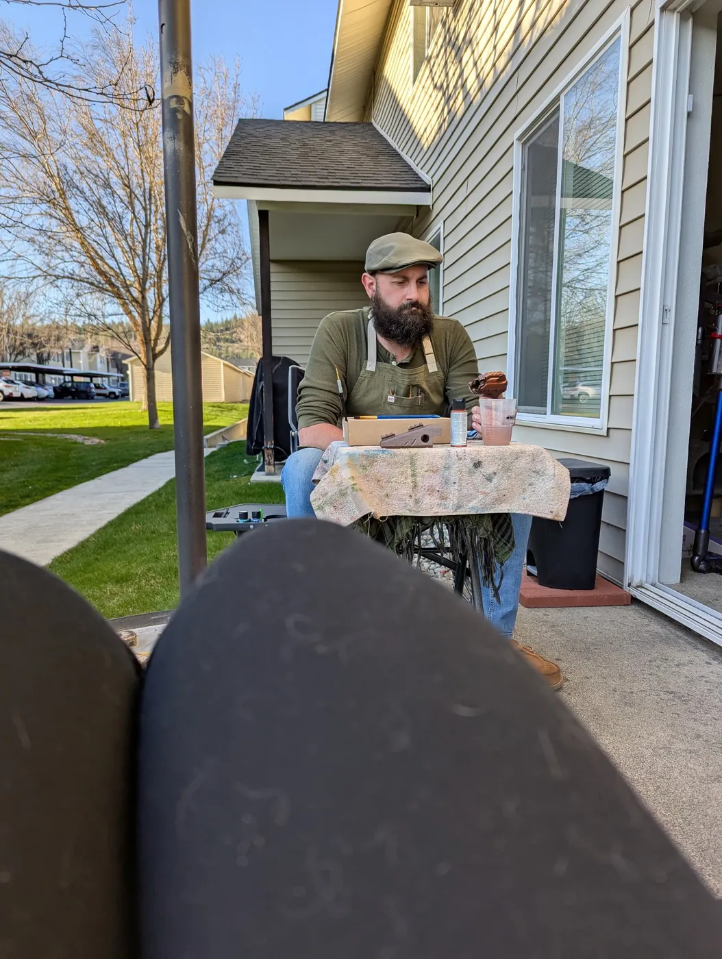 Looking out through the patio door at Tyler at the workbench in an olive shirt and cap, a lawn and the neighboring building behind him