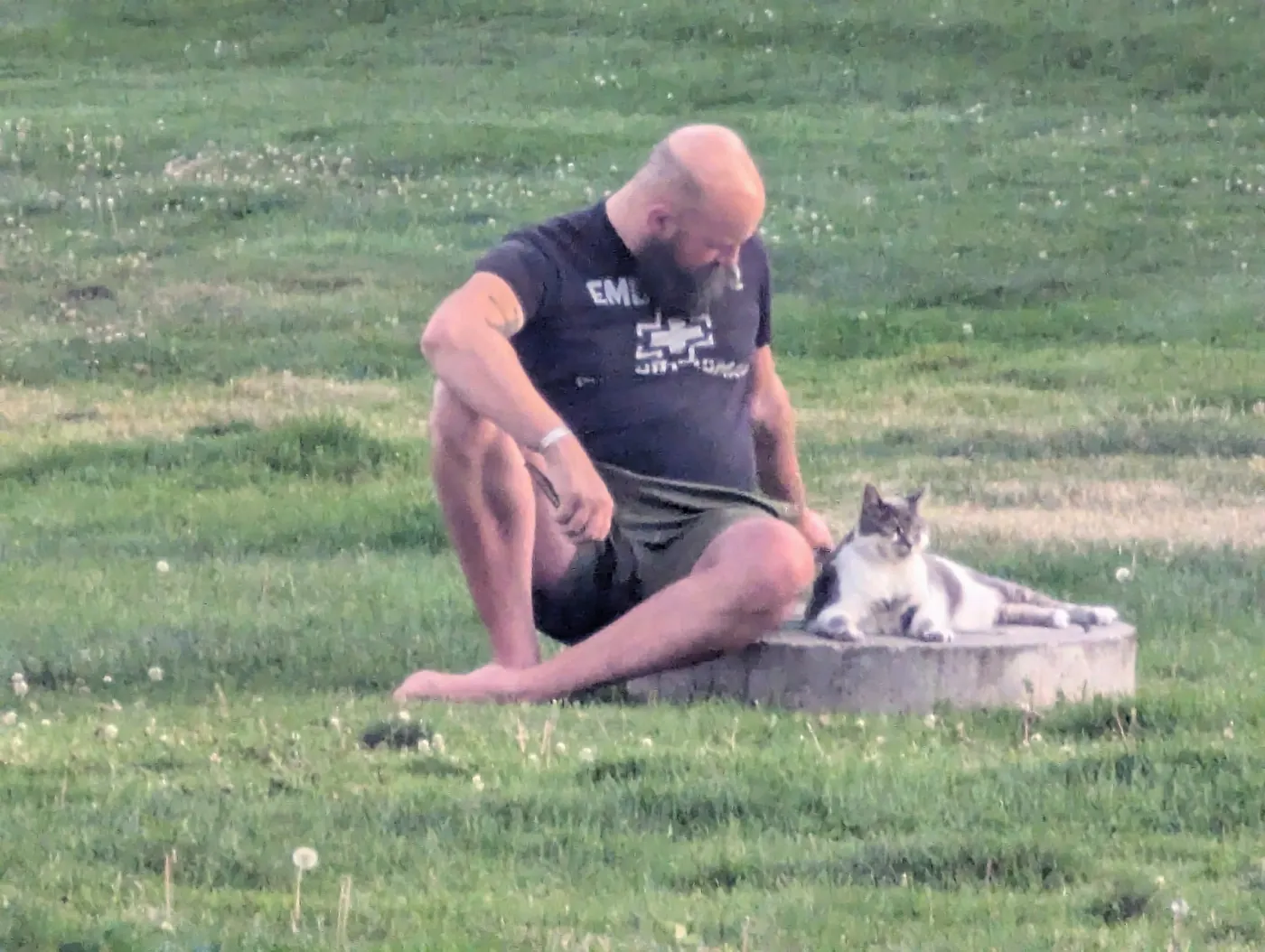 Tyler sitting on the lawn beside a tree stump where Gideon, a grey-and-white cat, is lying