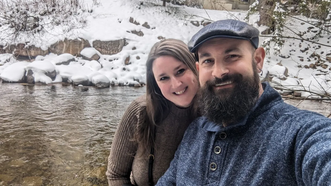 Selfie of Bethy and Tyler, cheek to cheek and smiling, in front of a snowy riverbank. He wears a flat cap and a blue sweater